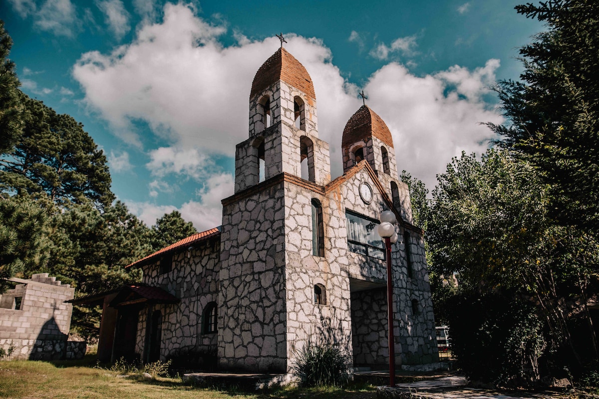 A stone building with twin towers and arched windows is featured under a partly cloudy sky. Surrounding greenery and scattered trees enhance the natural setting, while a pathway leads toward the entrance. The architecture reflects rustic charm, blending harmoniously with the environment.