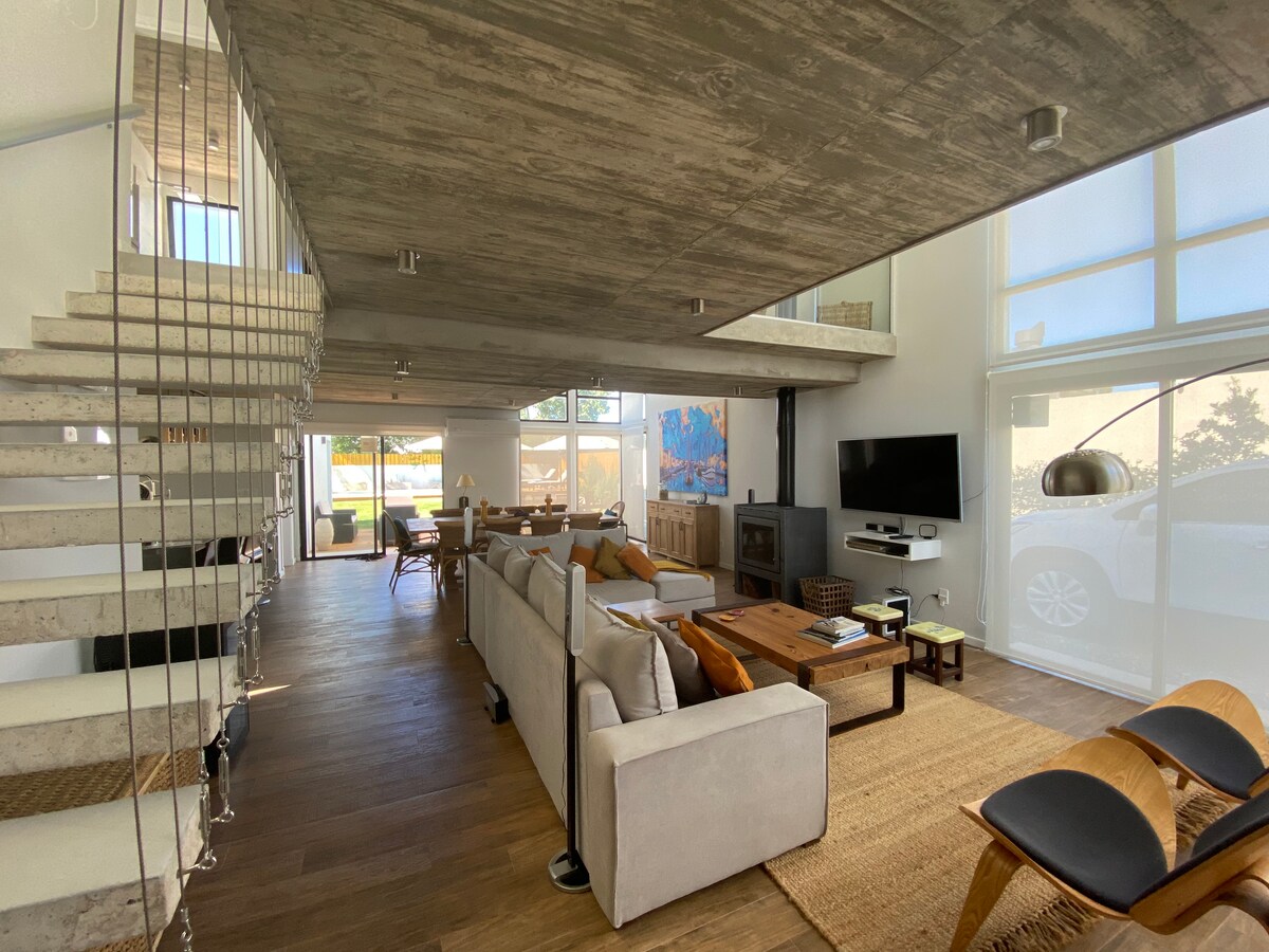 A spacious living and dining area is characterized by modern decor and natural light flooding through large windows. A comfortable seating arrangement is complemented by a wooden coffee table. A staircase leads to the upper level, and a TV is mounted on the wall, creating an inviting atmosphere.