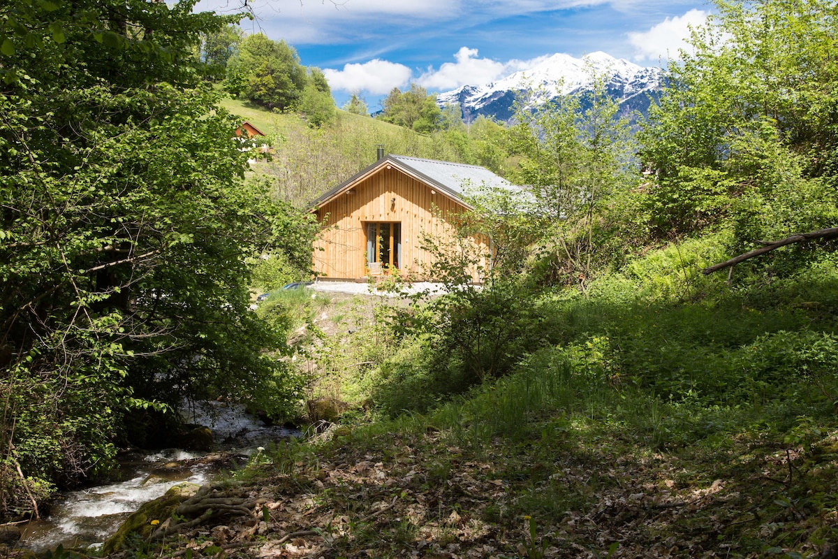A charming wooden chalet is nestled among lush greenery, with natural light highlighting its facade. A small stream flows nearby, complemented by mountainous scenery in the background. The structure is surrounded by trees, creating a serene, secluded atmosphere.