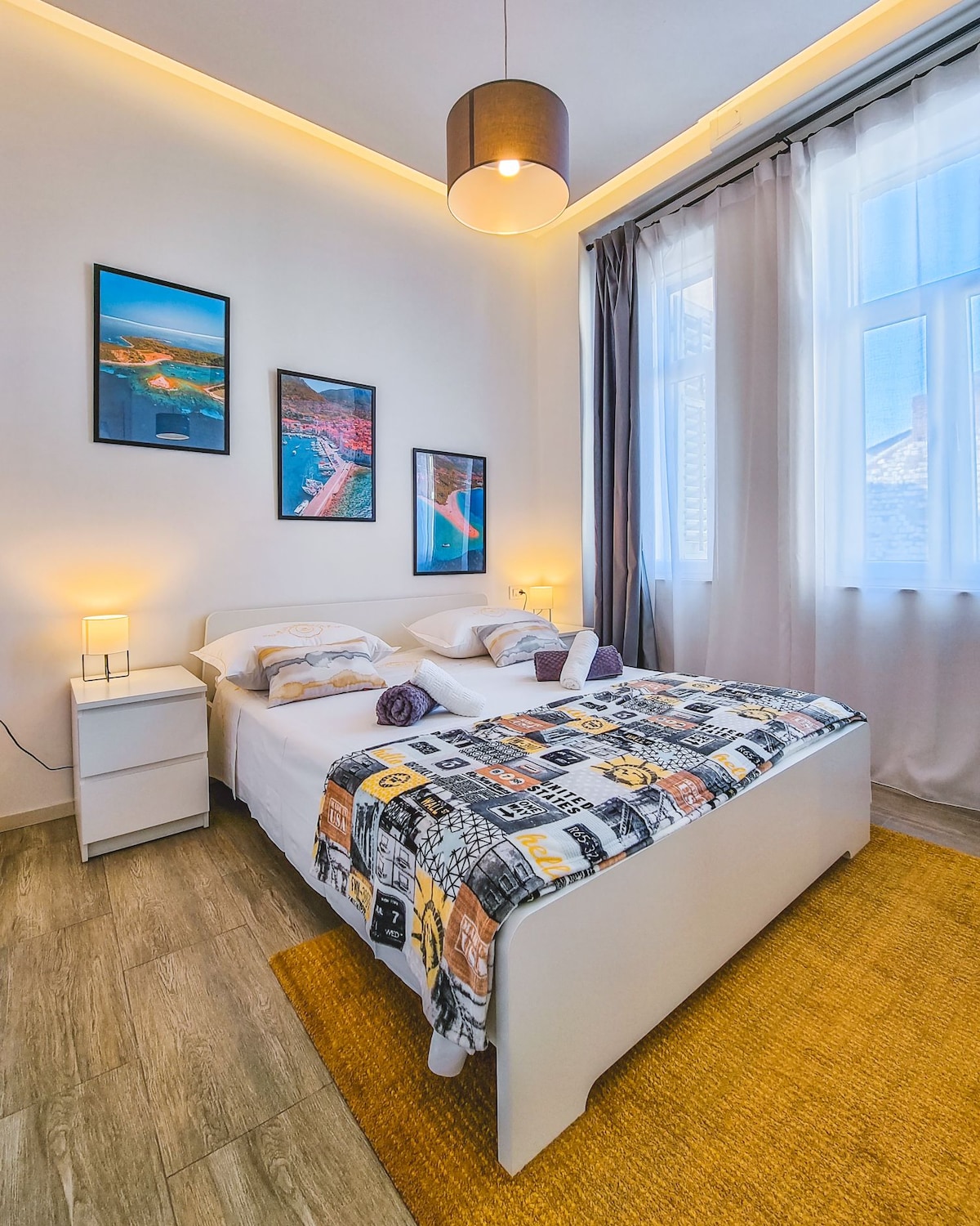 A cozy bedroom is showcased, featuring a spacious bed adorned with a patterned blanket. Two bedside lamps provide soft lighting beside the bed. Large windows with sheer curtains allow natural light to fill the room, while framed artwork adds visual interest to the walls.