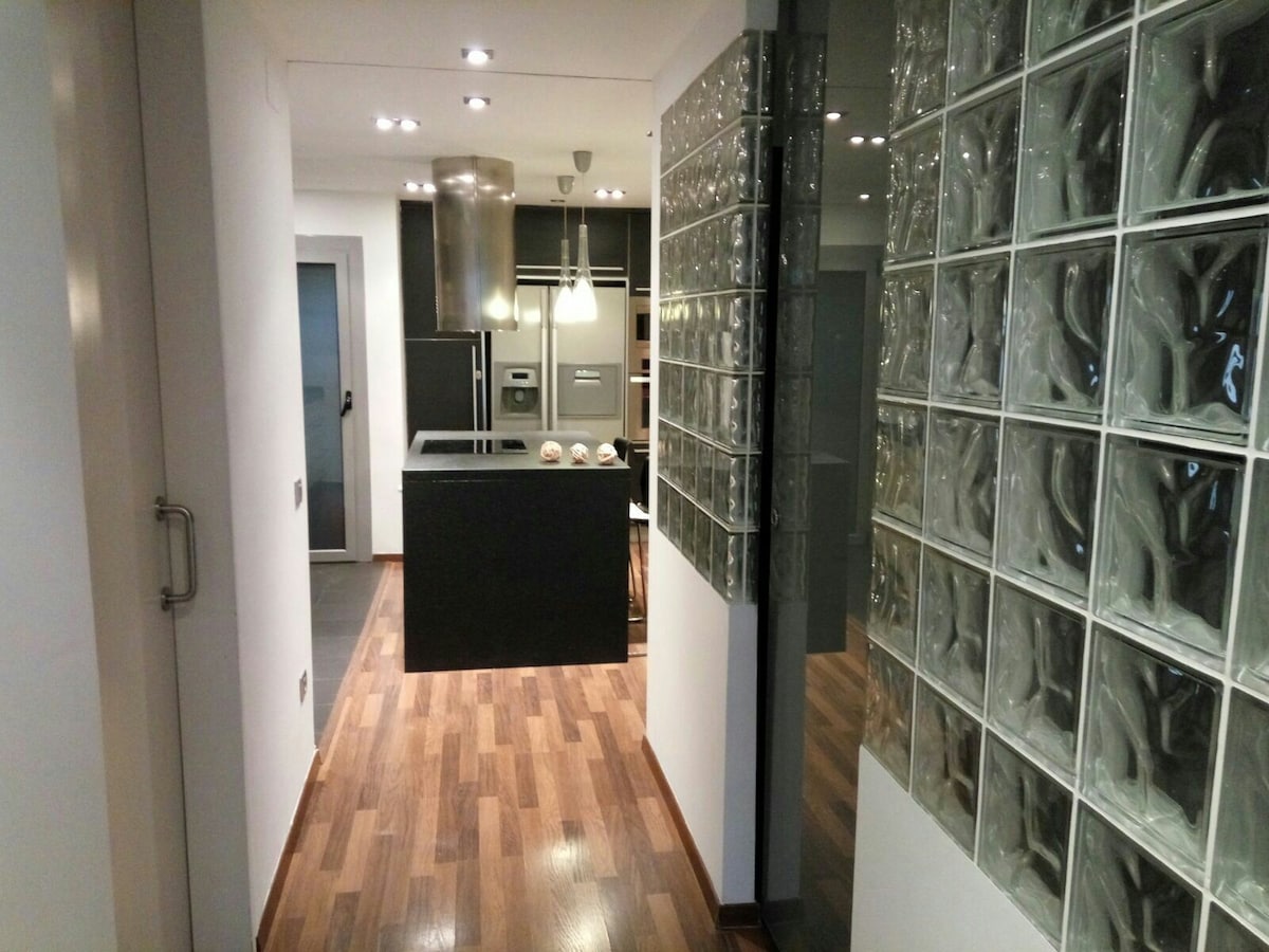 A modern hallway is viewed, featuring a glass block wall that allows light to filter through. The space leads to a sleek kitchen island with a dark countertop, surrounded by an inviting dining area. The warm wood flooring enhances the contemporary feel.