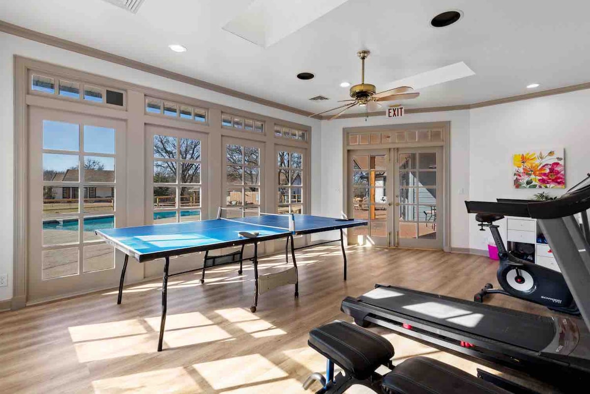 A bright activity area features two blue ping pong tables, surrounded by large windows that provide ample natural light. A treadmill is visible in the corner, and a ceiling fan hangs above, contributing to a comfortable environment.