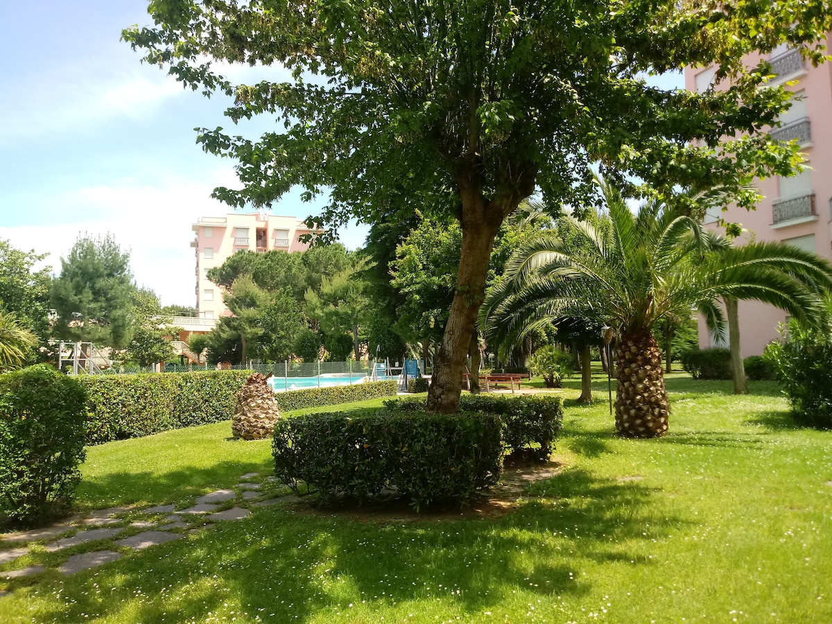 A well-maintained garden is visible, featuring lush green grass and several palm trees. The area is framed by neatly trimmed hedges and flower beds. In the background, a clear swimming pool is located within the residential complex, surrounded by trees and greenery.