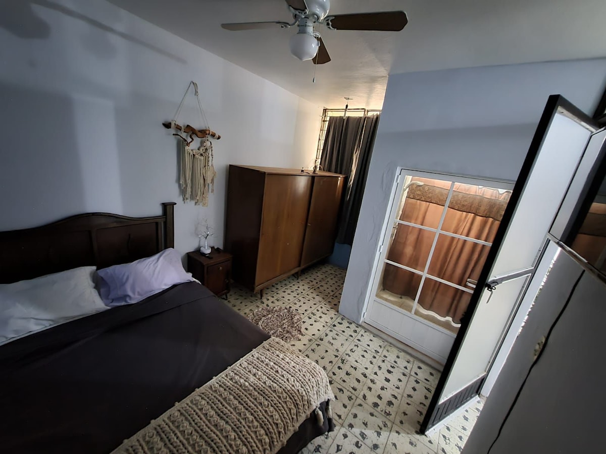 A bedroom featuring a spacious bed with neutral bedding, accompanied by a wooden nightstand and a wardrobe. Natural light enters through a door leading to a small outdoor area. The patterned flooring adds a subtle texture to the room.