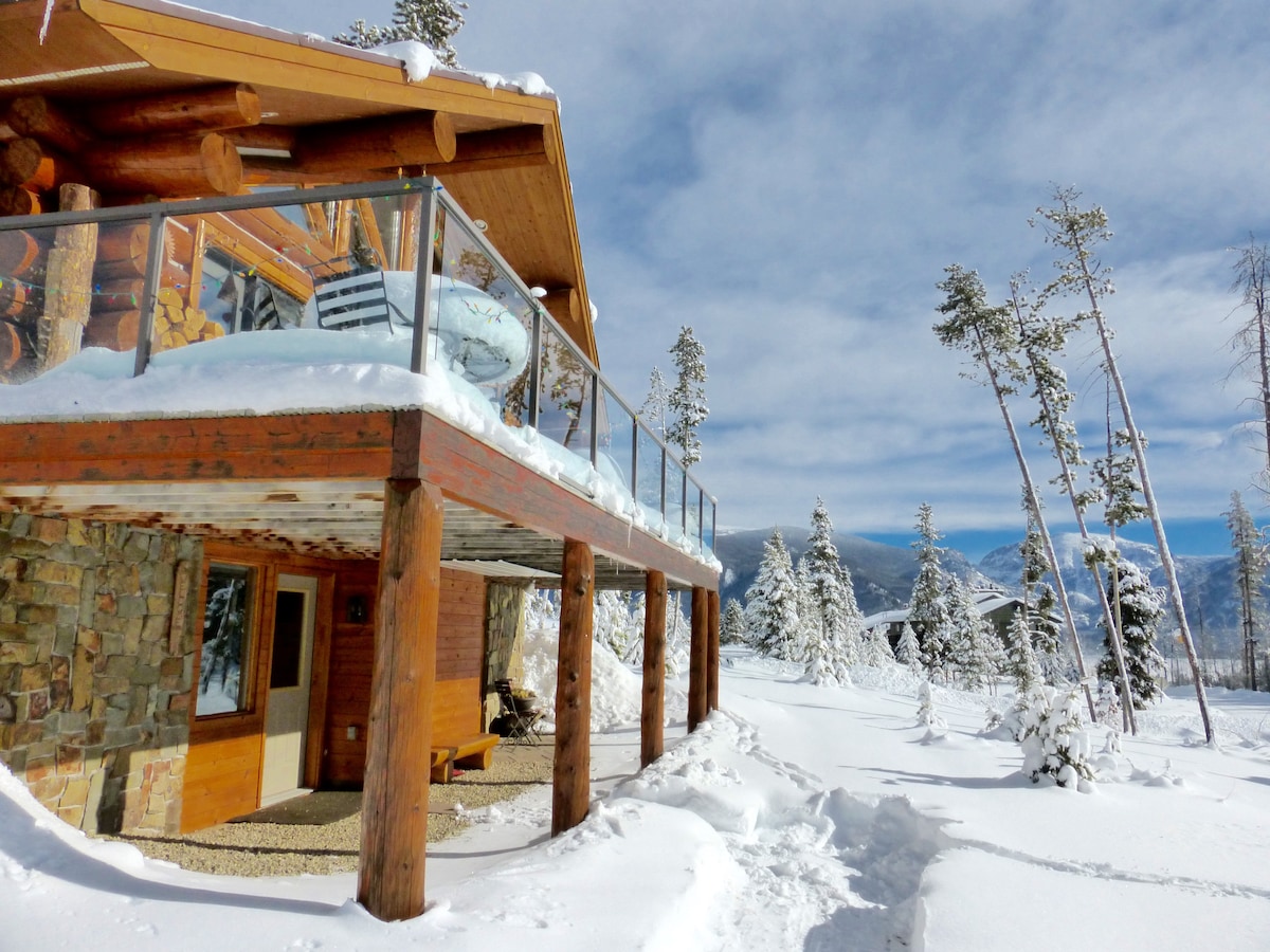 A snowy landscape surrounds the wooden structure, showcasing a balcony with glass railing. Snow-covered trees and mountains are visible in the backdrop, while a stone and wood entryway leads to a warmly designed suite.