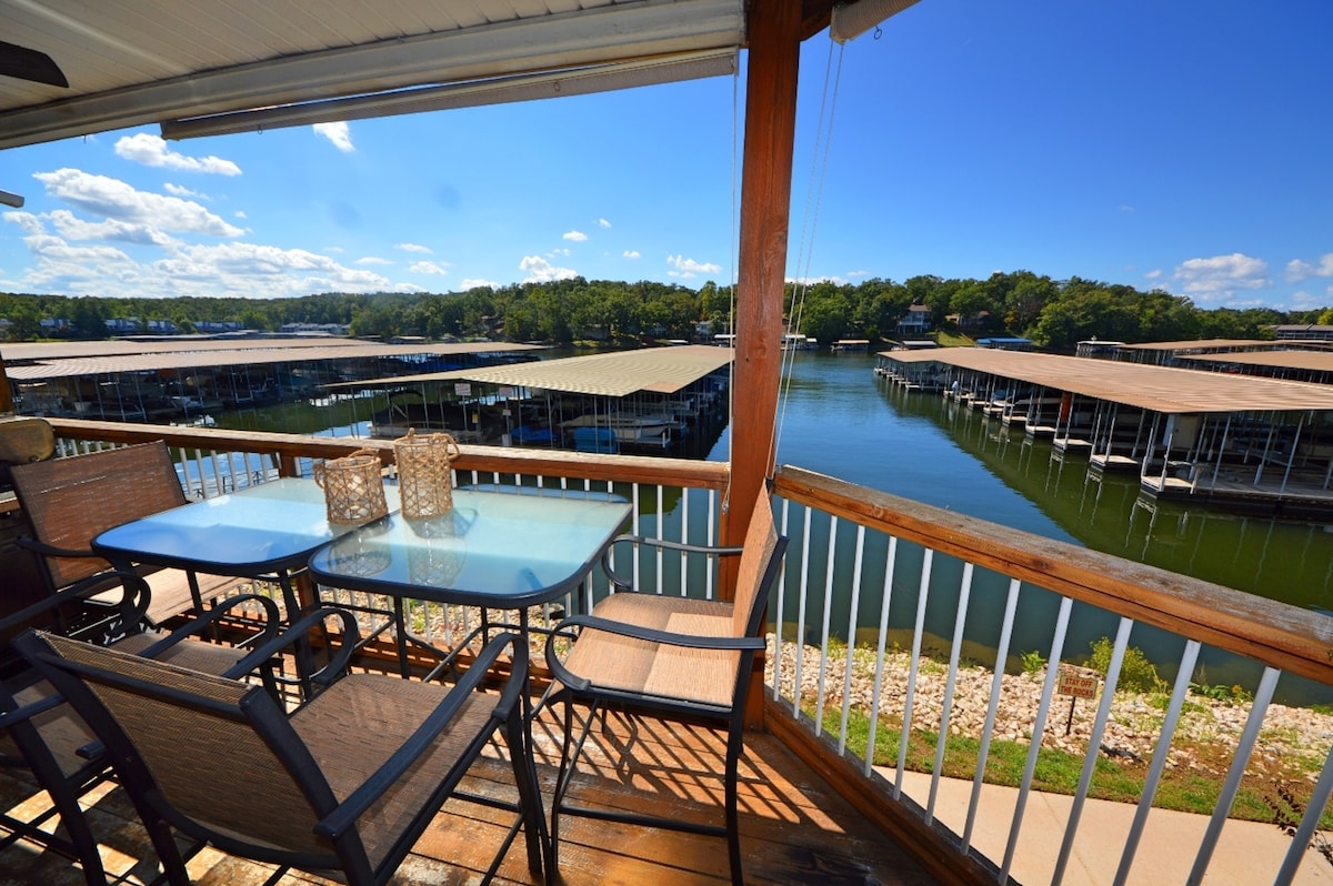 A deck area features a glass top table surrounded by chairs, overlooking a serene cove with multiple covered boat slips. Sunny skies and gentle clouds create a tranquil atmosphere, while the wooden railing provides safety and views of the water and surrounding trees.