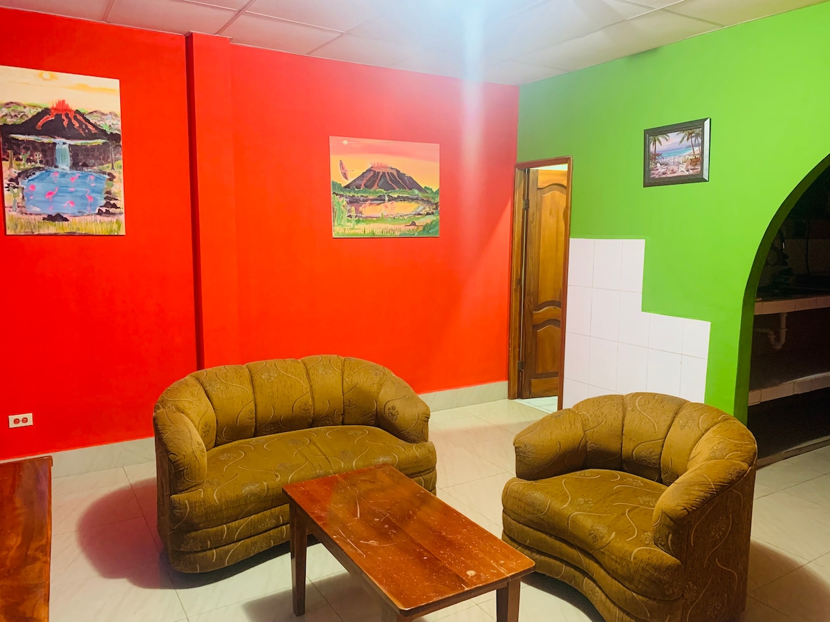 A living area features two circular, golden-brown armchairs positioned around a wooden coffee table. The walls are painted in vibrant red and green hues, complemented by framed landscape artwork depicting volcanic scenery. Light from overhead fixtures illuminates the space.