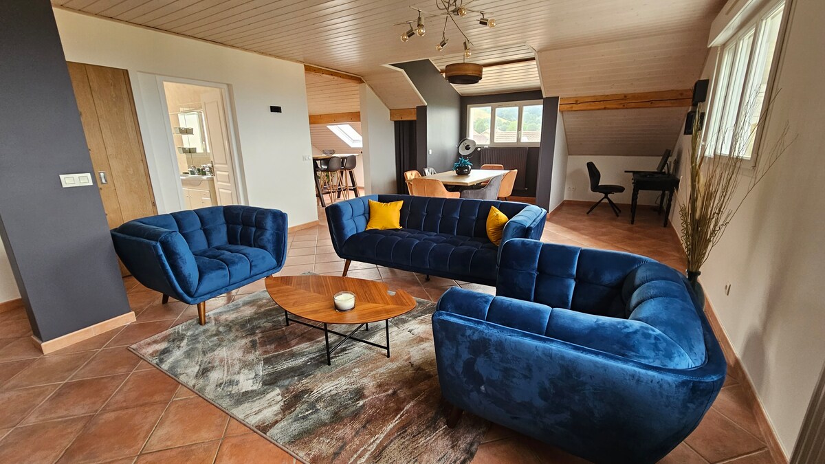 A spacious living area features modern blue velvet sofas and an oval coffee table. Natural light fills the room through large windows, illuminating the textured area rug. A dining area and workspace are positioned in the background, creating a functional and inviting environment.