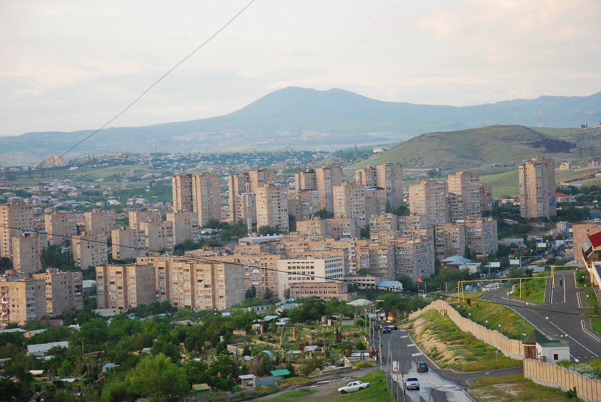 A sweeping view of an urban landscape is presented, featuring several tall residential buildings arranged in a grid. In the background, a mountainous terrain complements the cityscape, while green patches and roads intersect the scene.
