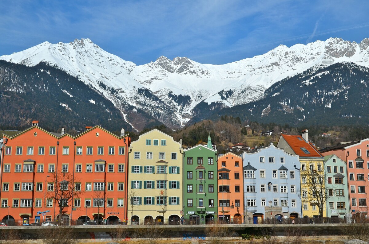 An array of colorful historic buildings lines the foreground, characterized by hues of orange, green, and blue. The majestic snow-capped mountains rise dramatically in the background, under a clear blue sky, creating a scenic and serene landscape.
