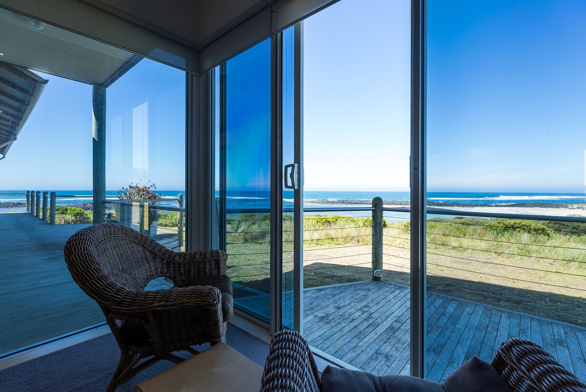 A comfortable seating area is framed by large glass doors, offering an unobstructed view of the Southern Ocean. The spacious outdoor deck can be seen beyond the grassy landscape, enhancing the connection between indoor and outdoor settings.