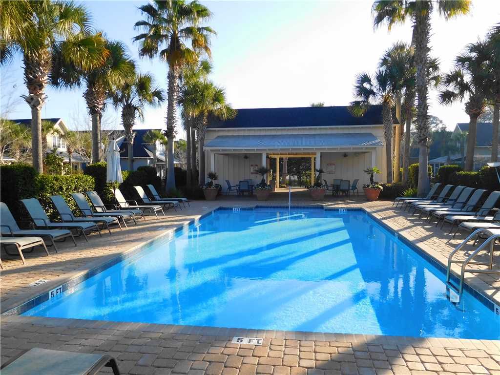 A spacious pool area is surrounded by lounge chairs, featuring palm trees that provide shade. The clear water is illuminated by sunlight, and a building with large windows is visible in the background, creating a relaxed atmosphere for guests.