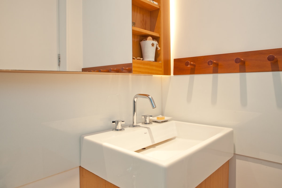 A modern bathroom sink area is presented with a sleek white basin. Wooden cabinetry is visible above, displaying neatly arranged decorative items. Two water faucets complement the clean lines of the design, with soft lighting enhancing the space.