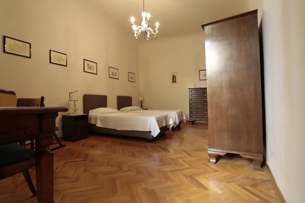 A spacious bedroom features two individual beds positioned next to each other. Wooden furniture includes a vintage wardrobe and a chest of drawers. A chandelier hangs from the ceiling, and framed artworks adorn the walls, enhancing the room's calm ambiance.