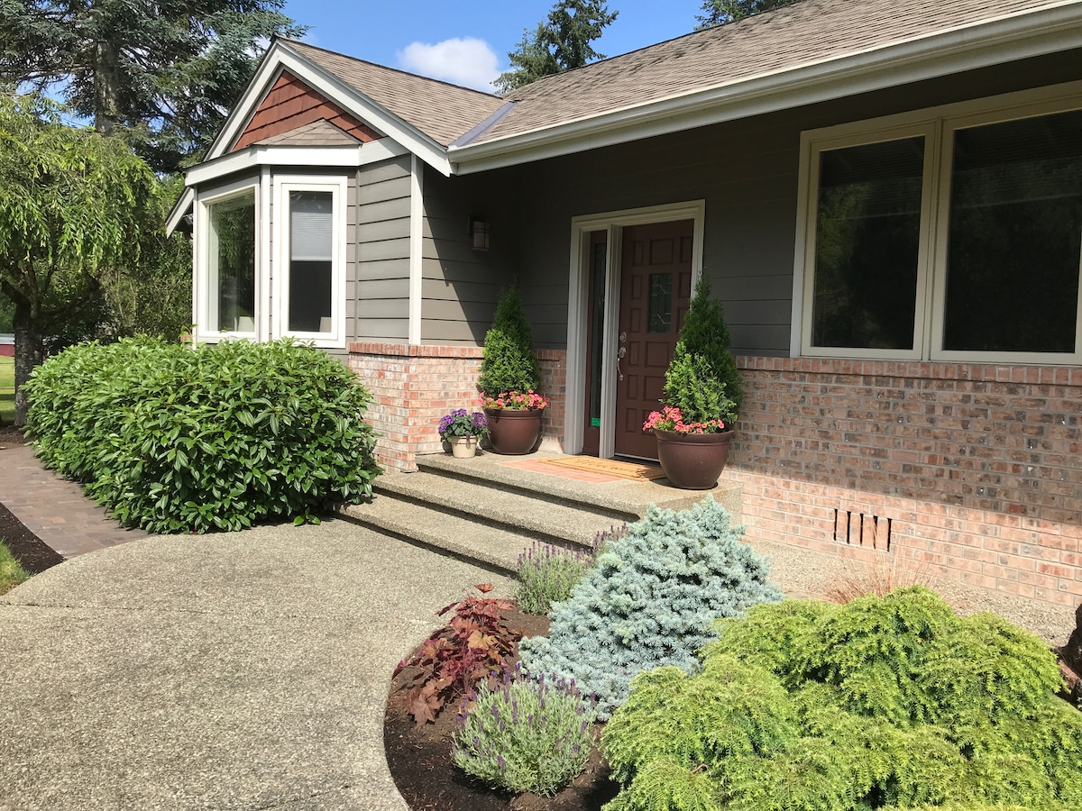 A welcoming entrance features a well-maintained path leading to the front door. Flanking the entrance are colorful flower pots, cultivated shrubs, and vibrant greenery, creating a pleasant introduction to the home. Large windows allow natural light to illuminate the front facade.