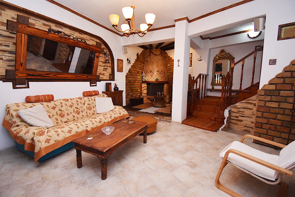 A spacious living area features a floral-patterned sofa and a wooden coffee table. An ornate mirror is mounted on the wall, reflecting the staircase leading to the upper floor. Natural light illuminates the room, creating a cozy atmosphere with stone and wood accents.
