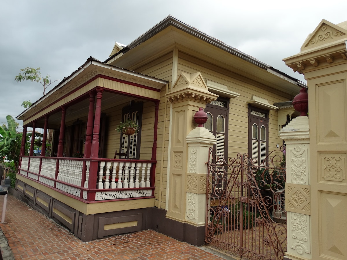 The exterior of the historic building showcases decorative wooden architecture with intricate details. A charming porch with red railings leads to the entrance, framed by ornate columns. An iron gate adds a classic touch to the well-maintained surroundings, inviting guests to explore the property.
