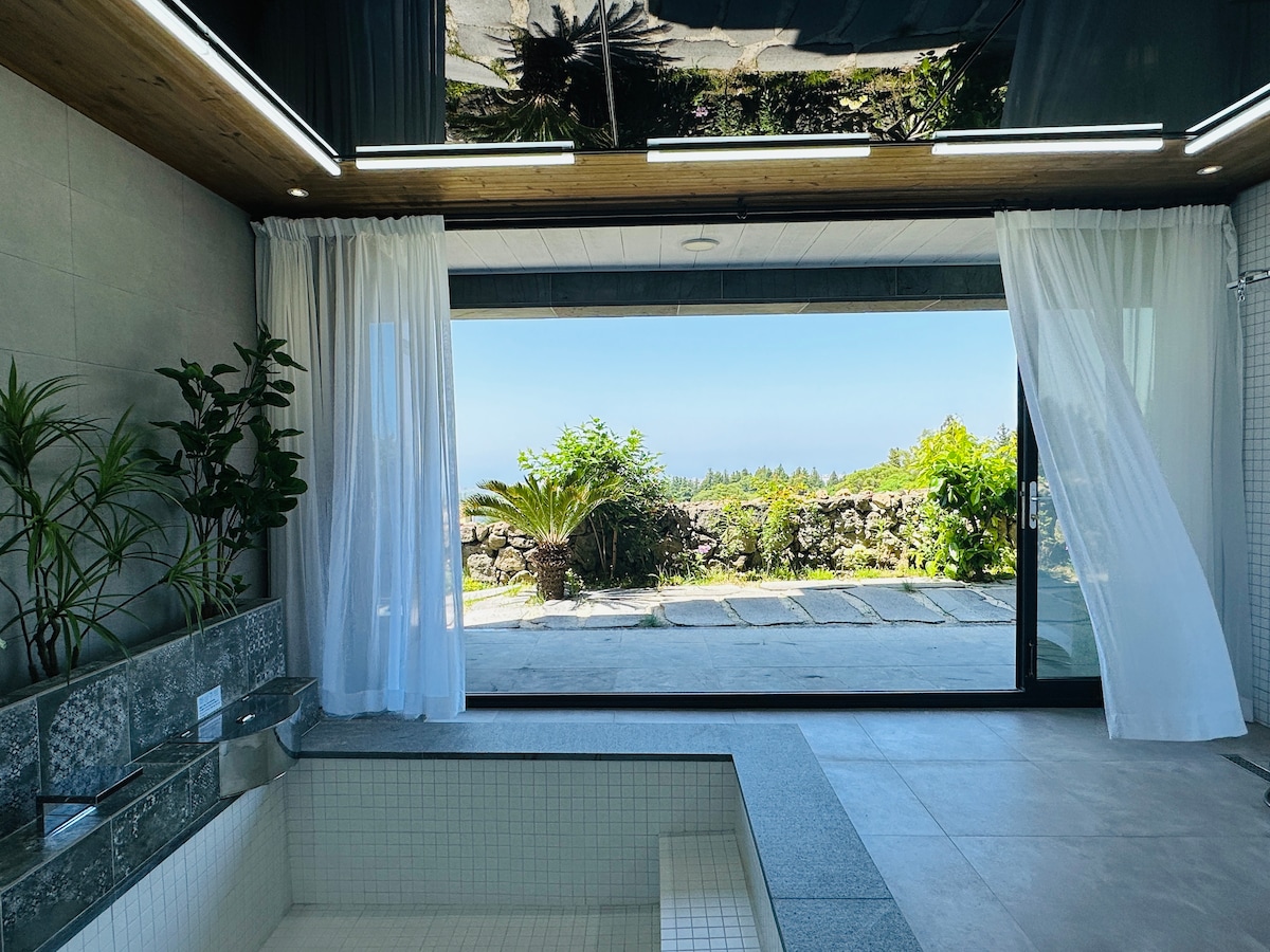 An indoor hot tub area features floor-to-ceiling glass doors that open to a garden view. Light, flowing curtains frame the entrance, and plants are placed along the walls, creating a refreshing atmosphere. The tub is situated on tiled flooring, enhancing a serene environment.