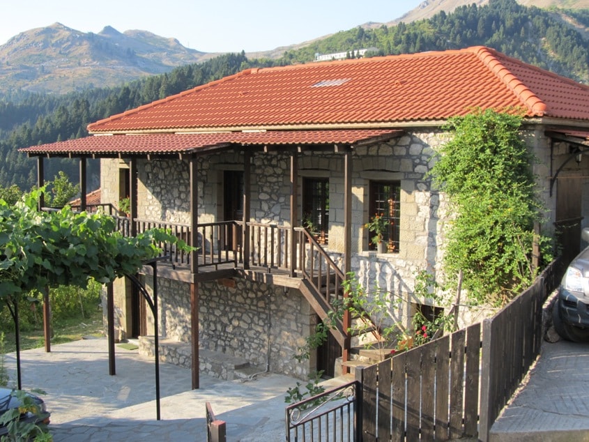 A traditional stone house is surrounded by lush greenery and mountain views. Wooden balconies and railings complement the rustic architecture. A paved pathway leads to the entrance, while nearby trees provide shade. The red-tiled roof adds charm to the tranquil setting.