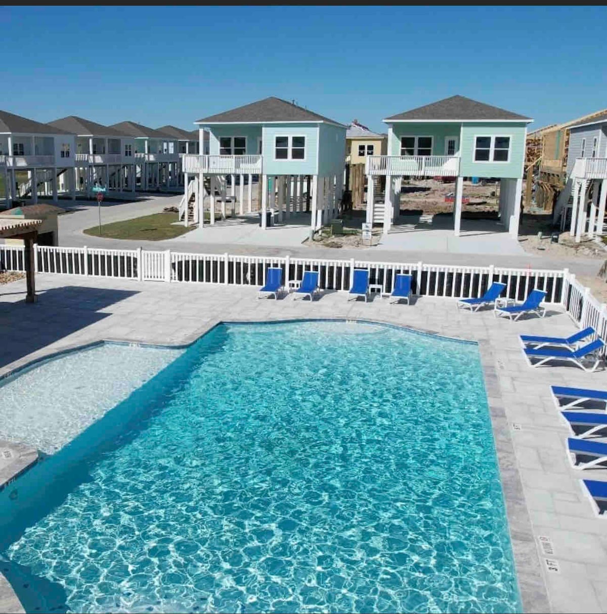 A community pool is featured in the foreground, surrounded by several lounge chairs. In the background, elevated coastal homes with light green exteriors are visible against a clear blue sky. The pool's water reflects shimmering light on the tile surface.