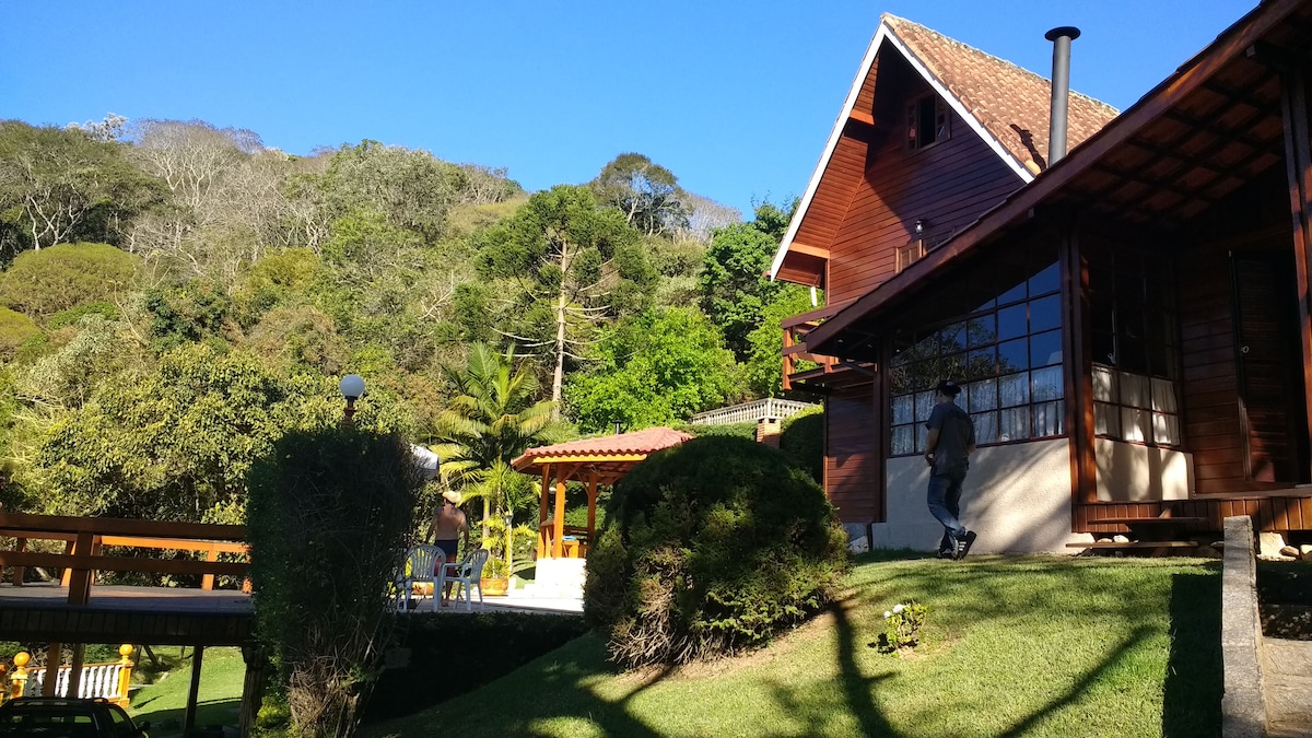 A wooden chalet is framed by lush greenery, showcasing a harmonious blend of nature and architecture. The structure features large windows and a sloped roof, while a nearby terrace is highlighted by outdoor seating. Tall trees and shrubs contribute to the scenic environment.