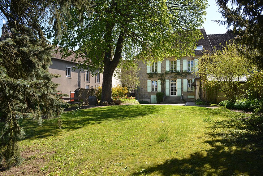 A spacious garden is framed by mature trees, providing ample greenery. A two-story house with several windows and a welcoming entrance is visible in the background. Sunlight illuminates the lawn, enhancing the serene outdoor space.