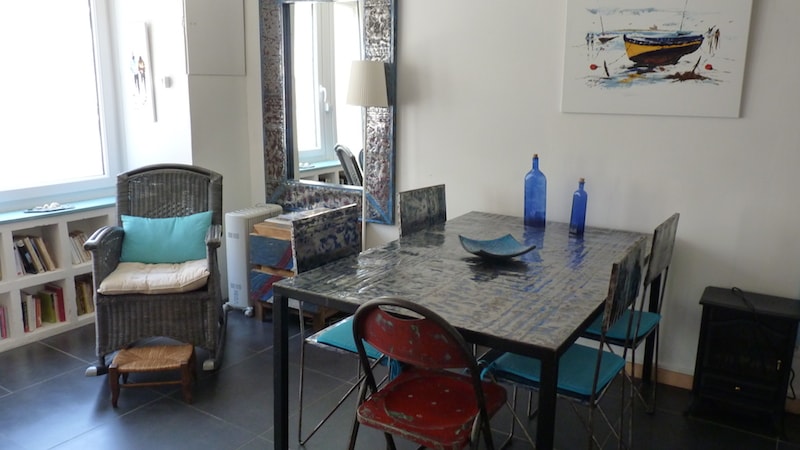 A bright dining area features a large table surrounded by six chairs. A wicker chair sits nearby, complemented by a square mirror reflecting light. A colorful artwork is hung on the wall above the table, and decorative blue bottles are placed strategically for visual interest.