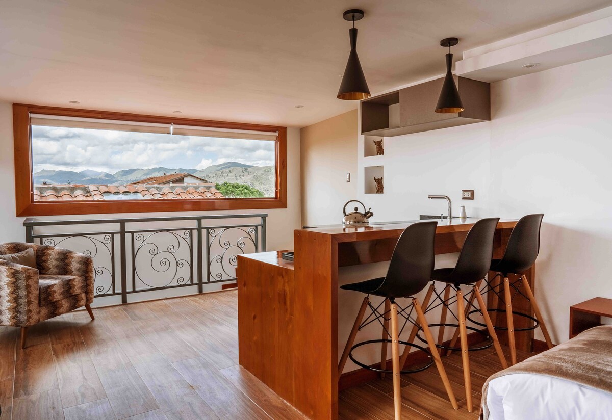 A bright and airy living space features a large window with mountain views. A wooden bar with three modern stools is positioned adjacent to the kitchenette. Cozy armchairs provide additional seating, while architectural details enhance the contemporary design.