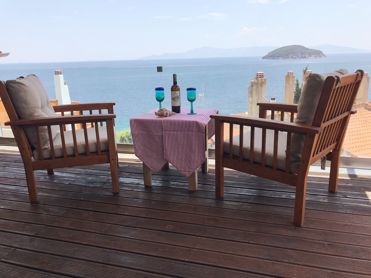 Two wooden chairs are positioned facing a stunning ocean view, accompanied by a small table draped with a striped cloth. On the table, two blue glasses and a bottle create an inviting setting, enhancing the tranquil atmosphere of the space.