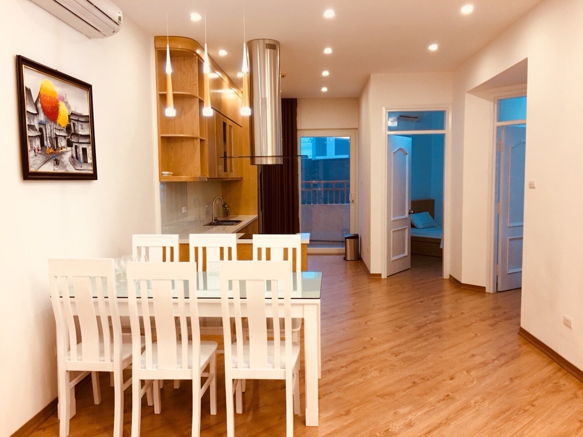 A modern dining area features a white dining table surrounded by six matching chairs. Natural light illuminates the wooden floor and walls, while a piece of artwork adorns the wall. The open layout connects to a well-equipped kitchen, visible in the background.