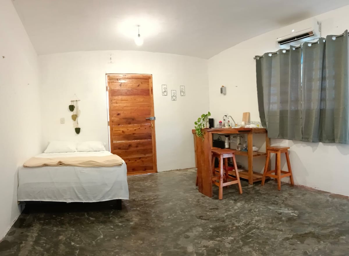 A simple room features a bed with neutral bedding and a wooden door. A small wooden table with two stools is positioned nearby. Large windows with dark curtains allow natural light to enter, creating a space that feels open and inviting.