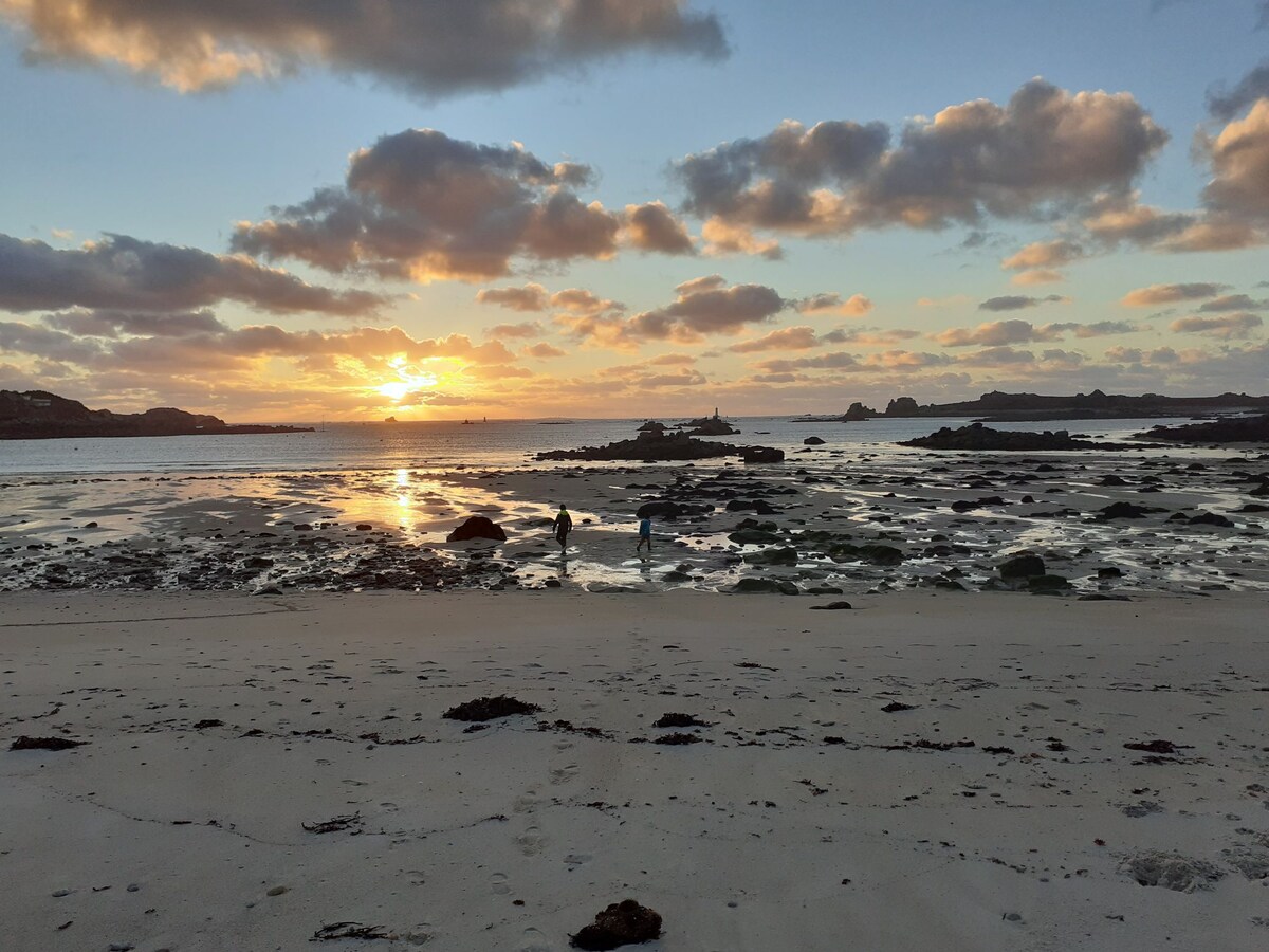 A tranquil beach scene is captured at sunset, with soft waves lapping at the shore. Silhouettes of individuals can be seen exploring the rocky coastline. The sky is painted with shades of orange and pink, reflecting upon the water's surface amidst scattered clouds.
