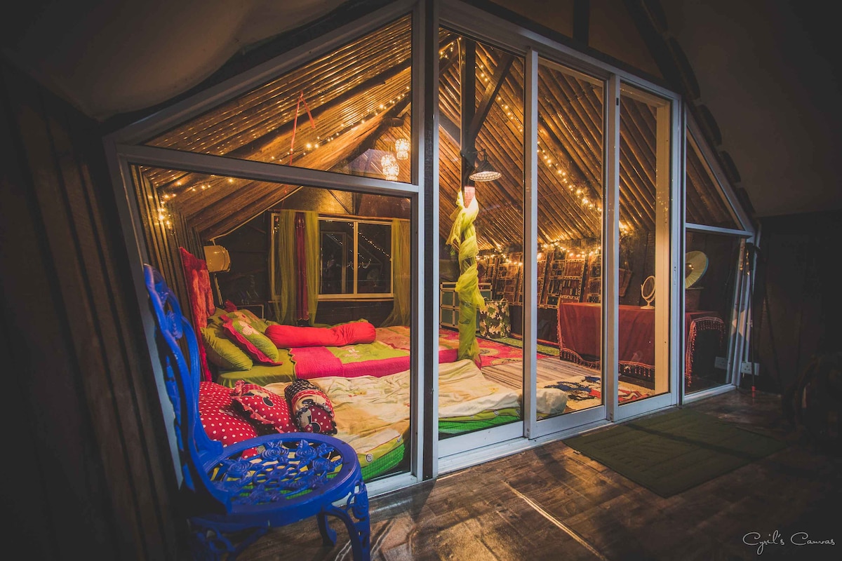 A spacious upper-level family room is shown, featuring large glass windows that offer a view of the surroundings. Warm lighting emanates from decorative string lights, casting a cozy glow over colorful cushions and a wooden table. Comfortable seating arrangements invite relaxation.