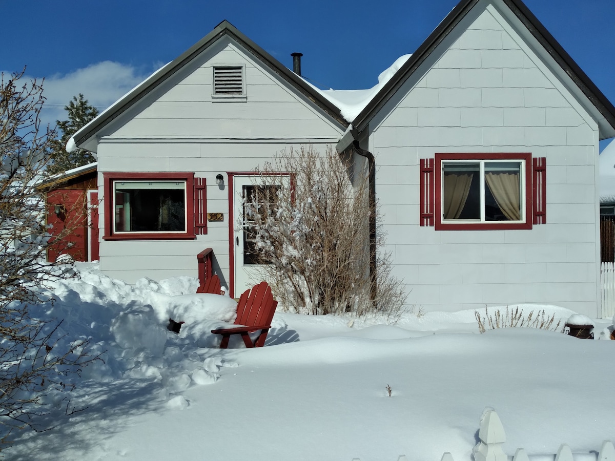 The exterior of a charming white house is shown, accented with red shutters. Snow blankets the ground and surrounds the building. Outside, two red chairs are positioned on the snow-covered yard, offering a cozy space to enjoy the winter scenery.
