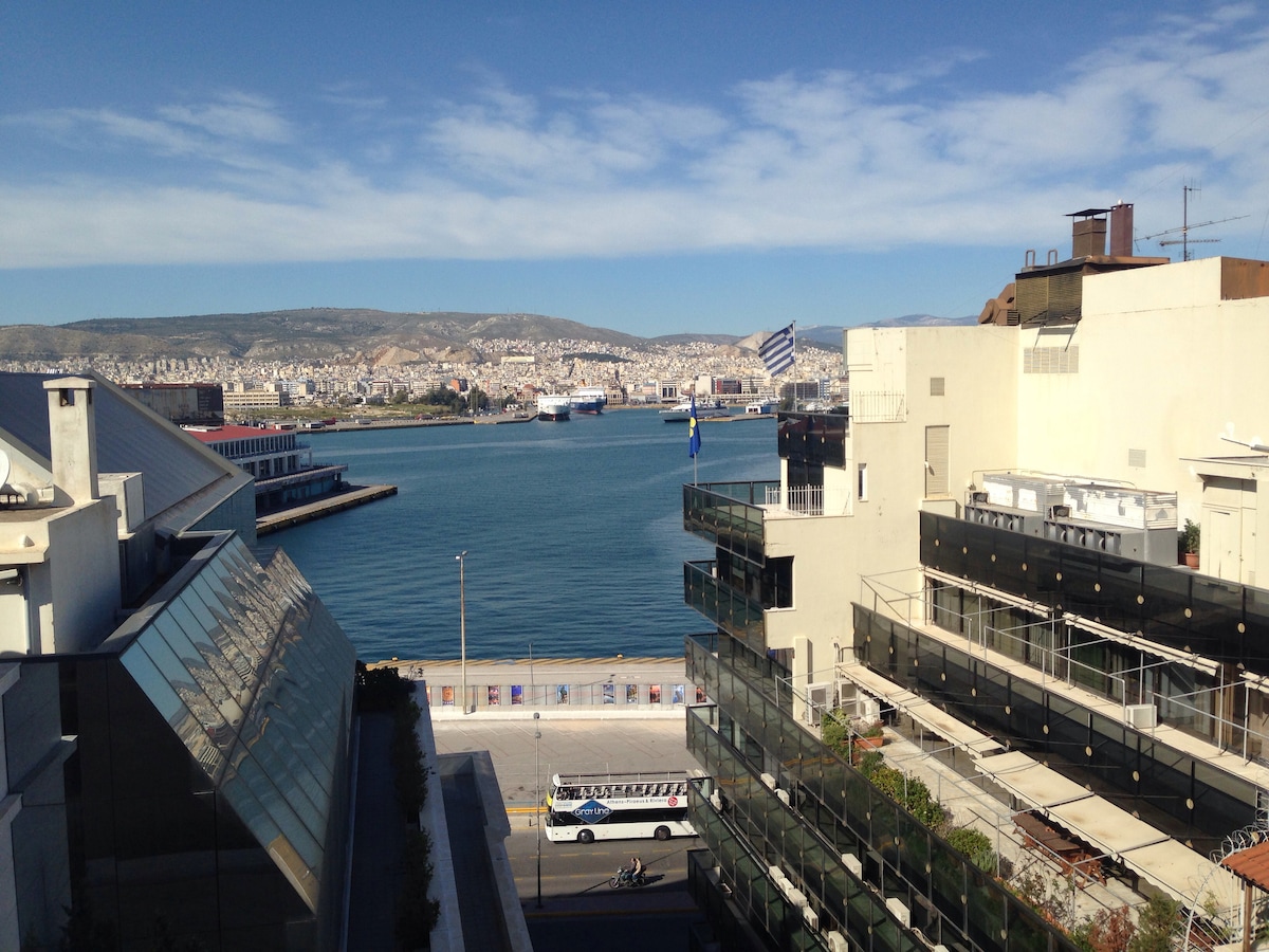 A view overlooking the tranquil waters of the port, framed by modern buildings. Several vessels can be seen in the distance, while the skyline features hills and a clear blue sky. The area appears well-maintained, suggesting a vibrant atmosphere.