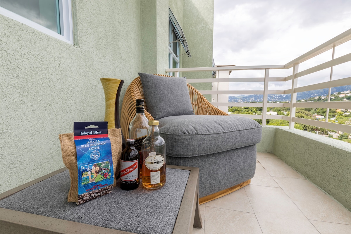 A comfortable seating area is displayed on a private balcony, featuring a grey cushioned chair alongside a small table. Several bottles of liquor and a snack bag are placed on the table, with a scenic view of the surrounding landscape visible in the background.