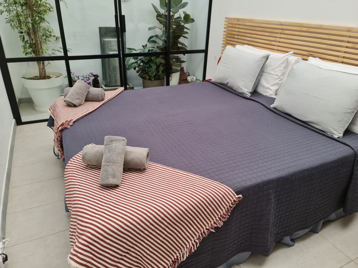 A spacious bed is presented with a dark-colored quilt and striped throws draping over the edges. Soft pillows are arranged at the headboard, which features a natural wood design. Large glass panels provide a view of indoor plants, enhancing the room's connection to nature.