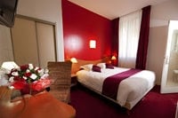 A well-appointed room features a spacious bed with decorative pillows against a bold red wall. A seating area with a cozy armchair and a small table is visible, alongside an arrangement of flowers. Natural light filters through a sheer window, illuminating the room.
