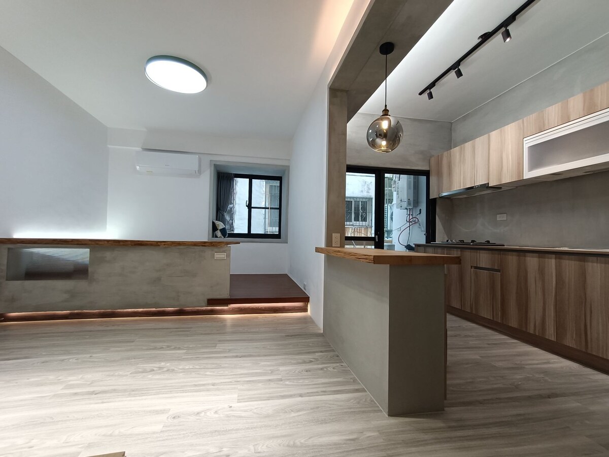 An open living space is depicted, featuring a modern kitchen with light wood cabinetry and a sleek countertop. Natural light enters through large windows, illuminating the minimalist design. A subtle illumination along the flooring highlights the spaciousness, with a seating area visible in the background.