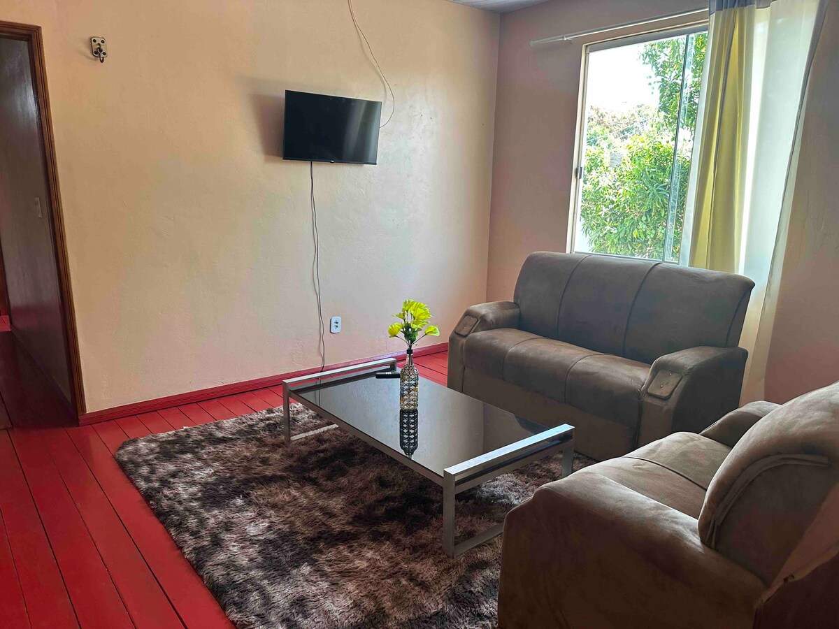 A spacious living area features two comfortable sofas arranged around a glass coffee table. A vibrant flower vase adds a touch of life to the table. Natural light streams in through a large window, complementing the warm tones of the wooden floor.