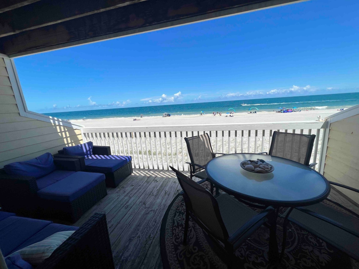 A private deck features a round dining table surrounded by chairs, offering direct views of the sandy beach and ocean. Comfortable lounge seating is positioned nearby. Bright blue skies and gentle waves complement this inviting outdoor space.