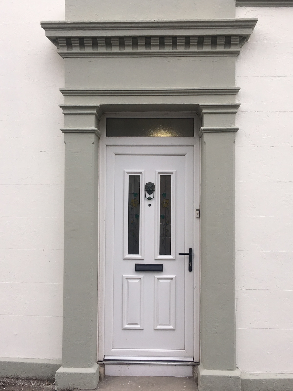 A white front door with a decorative panel is framed by a grey architrave. Three vertical glass panels adorned with frosted designs allow soft light to filter through. The door features a black handle and a letterbox, emphasizing its welcoming entrance.