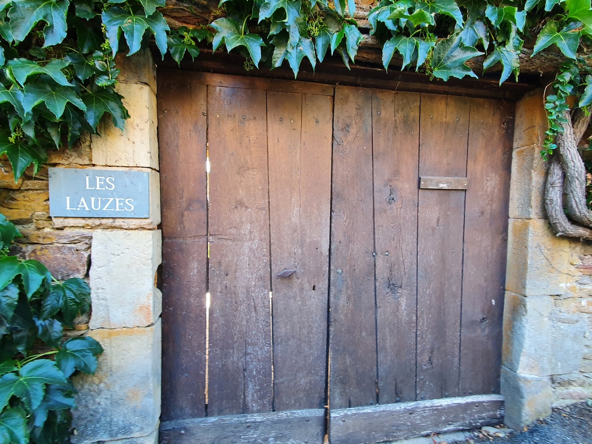 A rustic wooden door provides access to the property, framed by lush green ivy. A stone wall supports the door, with a sign labeled 'LES LAUZES' affixed nearby, enhancing the natural charm of the entrance.