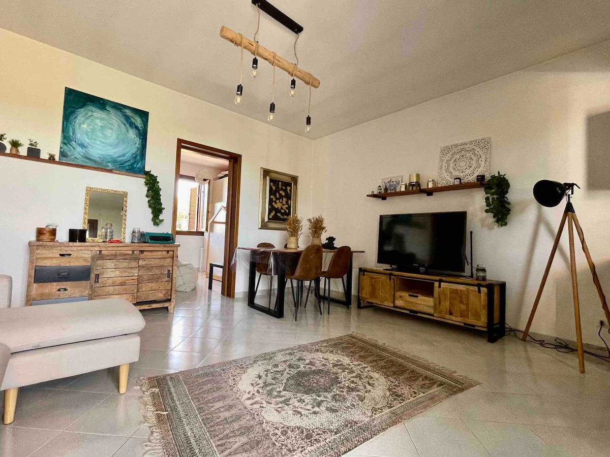 A well-decorated living area features a comfortable sofa, a stylish wooden sideboard, and a flat-screen TV. A dining table with chairs is placed beside a window, while decorative plants and artwork provide the finishing touches. Soft lighting enhances the room's welcoming ambiance.