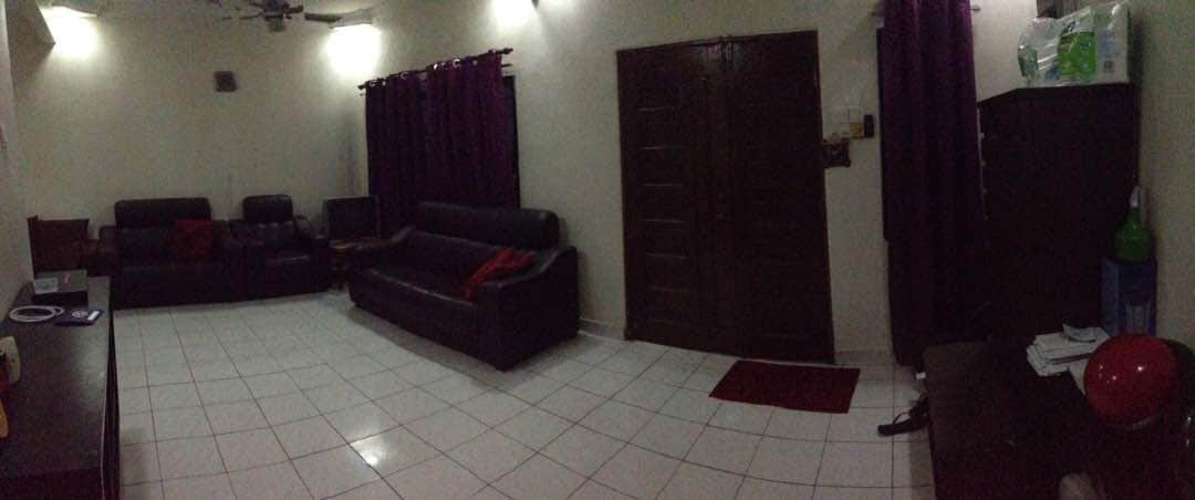 A spacious living area is presented with glossy white tile flooring and neutral-colored walls. Two black leather sofas are placed against the walls, with matching cushions. Dark curtains frame the windows, while a small side table and a wall-mounted cabinet are visible in the room.