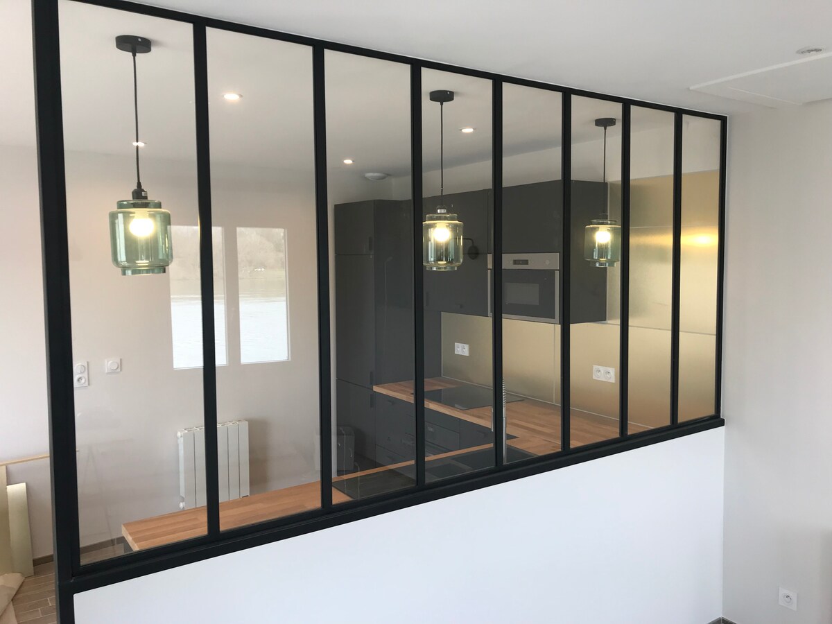 A modern kitchen is presented behind a glass partition, featuring sleek cabinetry and wooden countertops. Two pendant lights hang above the counter, illuminating the space. The reflection of natural light creates a bright atmosphere, while the design allows for an open view of the adjacent area.
