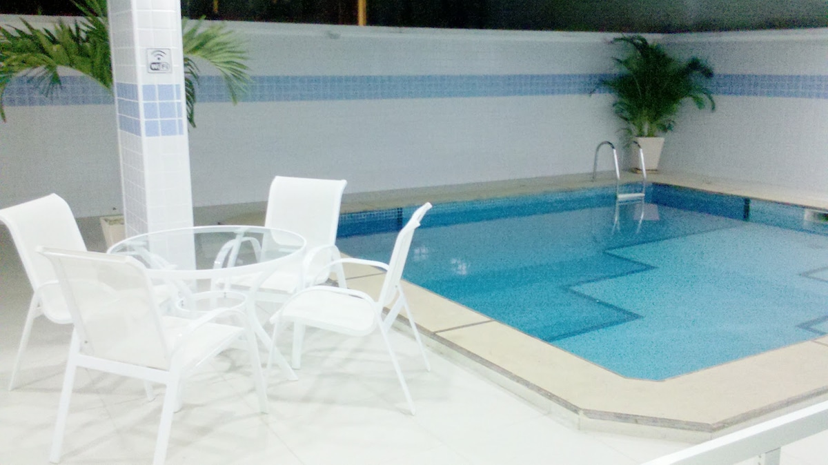 A private pool area is showcased with clear blue water, surrounded by white tiled walls. Two white lounge chairs and a round table are set beside the pool, with lush greenery visible in the background, offering a serene space for relaxation.
