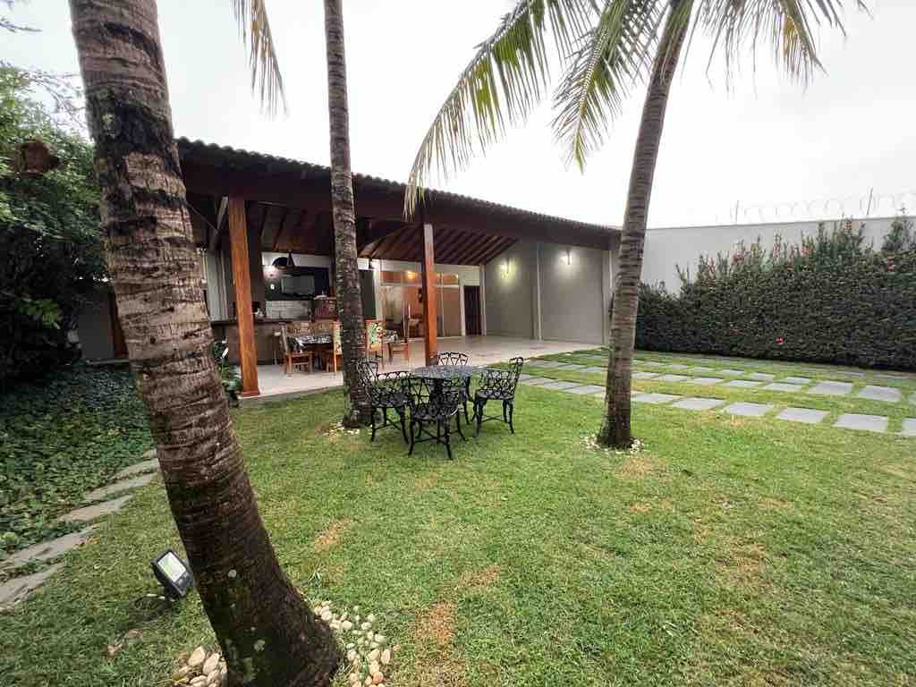 A spacious outdoor area features a well-maintained lawn with a dining set under a wooden pergola. Tall palm trees create a natural frame, while a barbecue grill is positioned nearby. Paved stones lead through the garden, enhancing the inviting ambiance for gatherings.