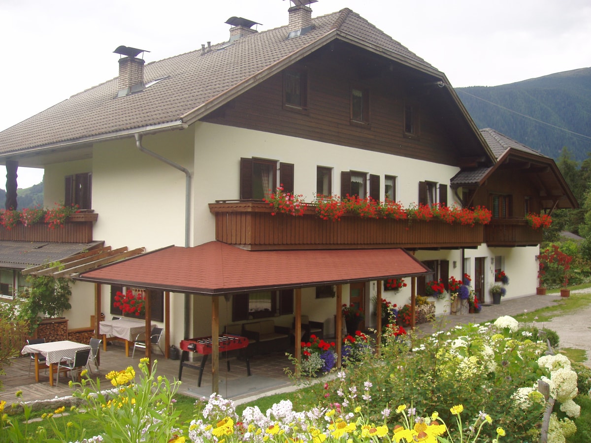 A two-story house is depicted, featuring a blend of wooden and stucco architecture. Flower boxes overflow with vibrant blooms along the balconies. An outdoor terrace adorned with tables and chairs invites relaxation, while a lush garden in the foreground adds to the serene atmosphere.