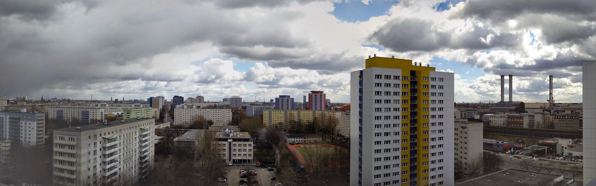 A panoramic view of the Berlin skyline is captured from the 10th floor, showcasing varied architectural styles. Residential buildings, sports facilities, and a distant industrial area are visible under a cloudy sky, providing a sense of the city's urban landscape.